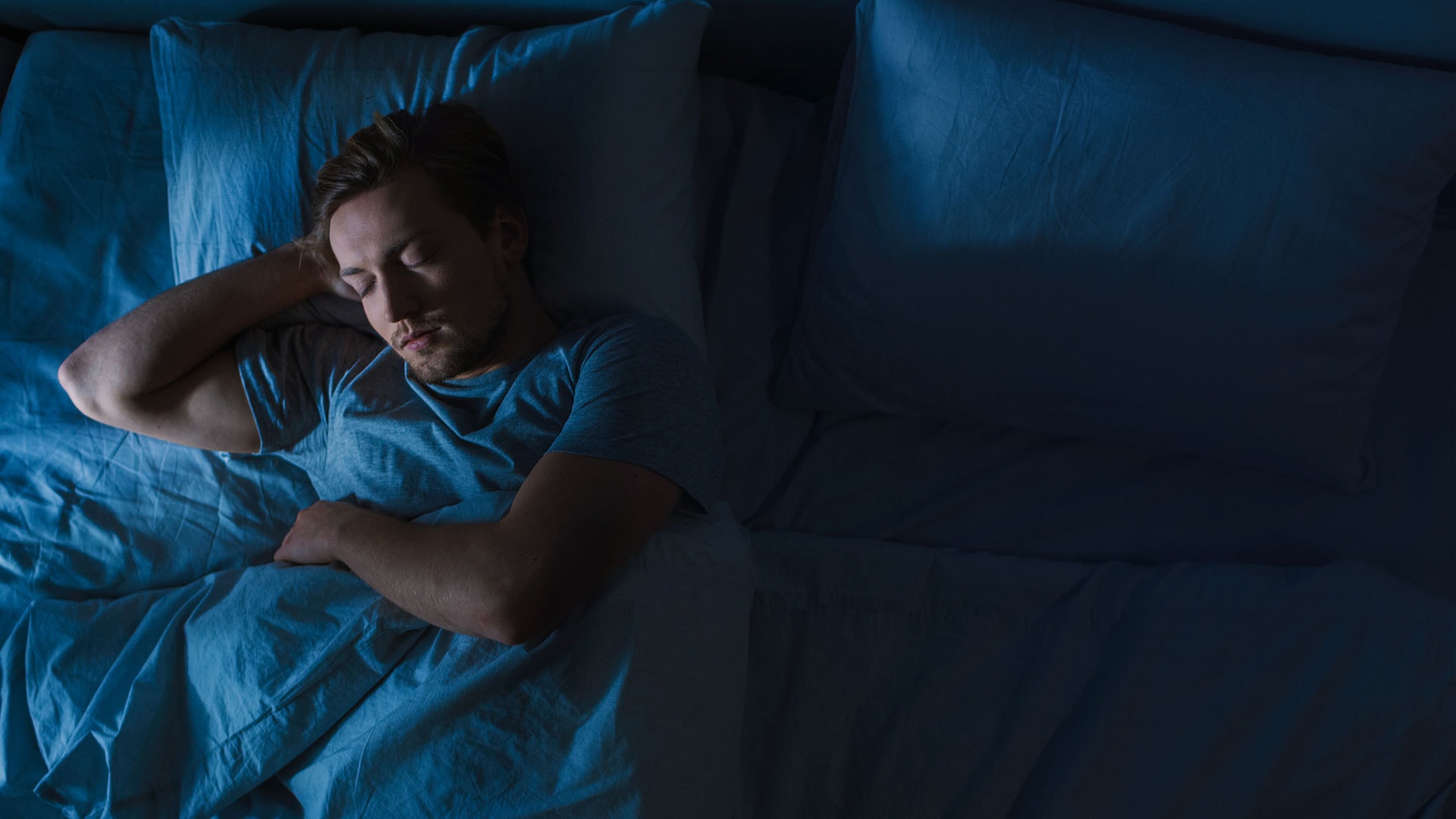 A white male with a short beard sleeps in a dark bedroom