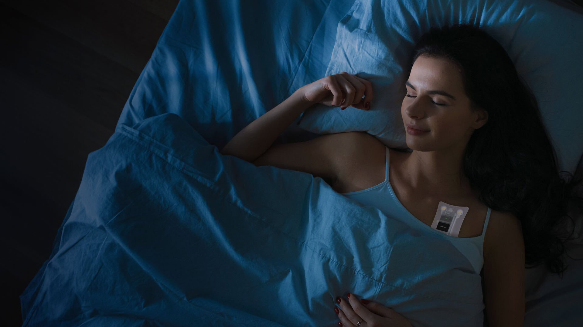A white women with long dark hair sleeps under blue sheers while wearing a Telepatch device